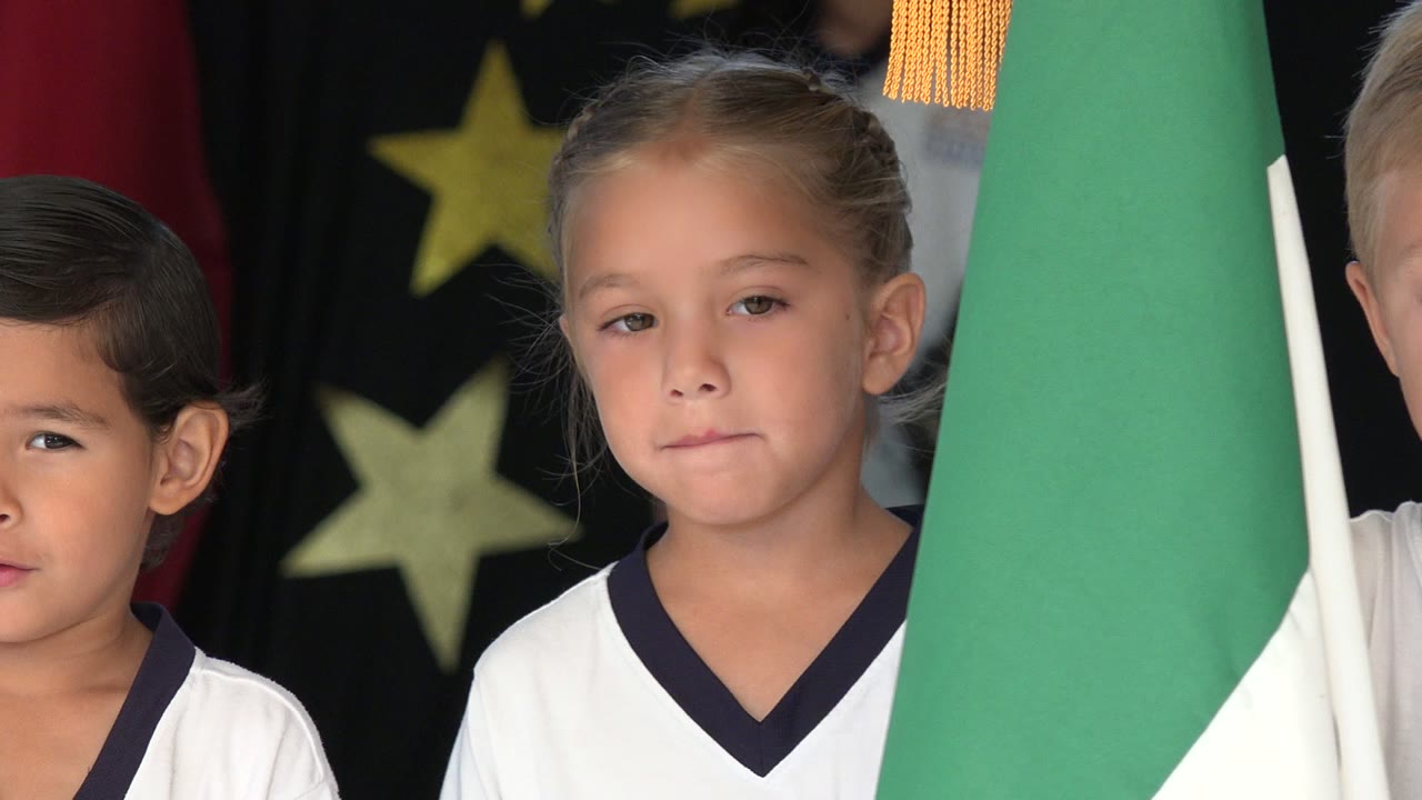 A girl in a white school uniform faces forward while a green and white flag fills the right side of the frame in San Jose del Cabo, Mexico.