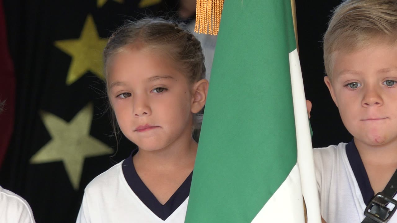 Girl in uniform stands near a flag with another child partly visible.