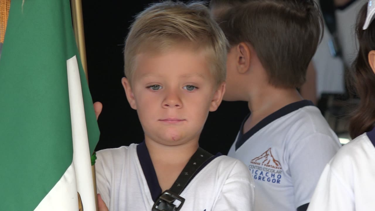 A blond young boy in a white shirt holds a green and white flag while other children stand beside him during a kindergarten graduation in San Jose del Cabo, Mexico.