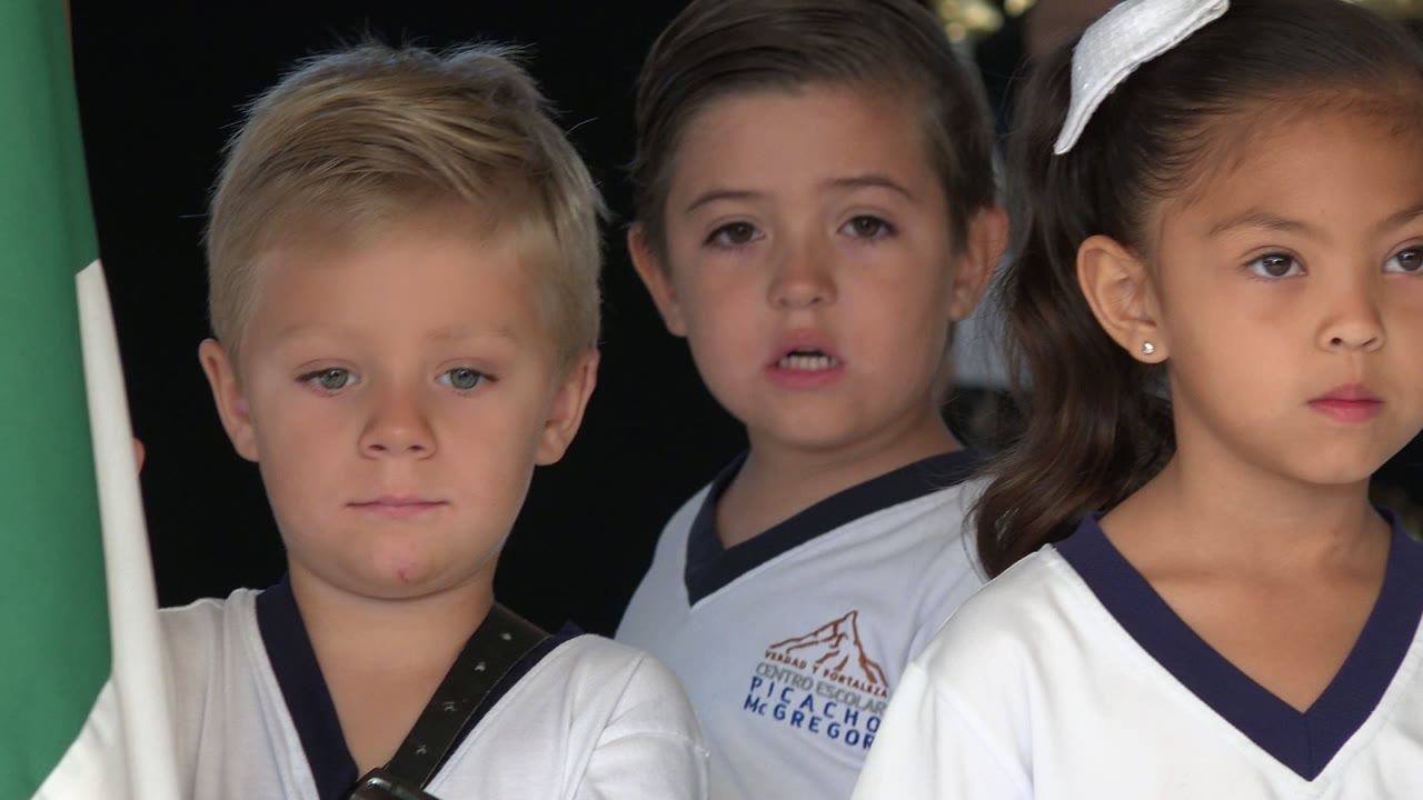 Three young children in matching white shirts stand shoulder to shoulder, with neutral faces and a dark ceremony backdrop in San Jose del Cabo, Mexico.
