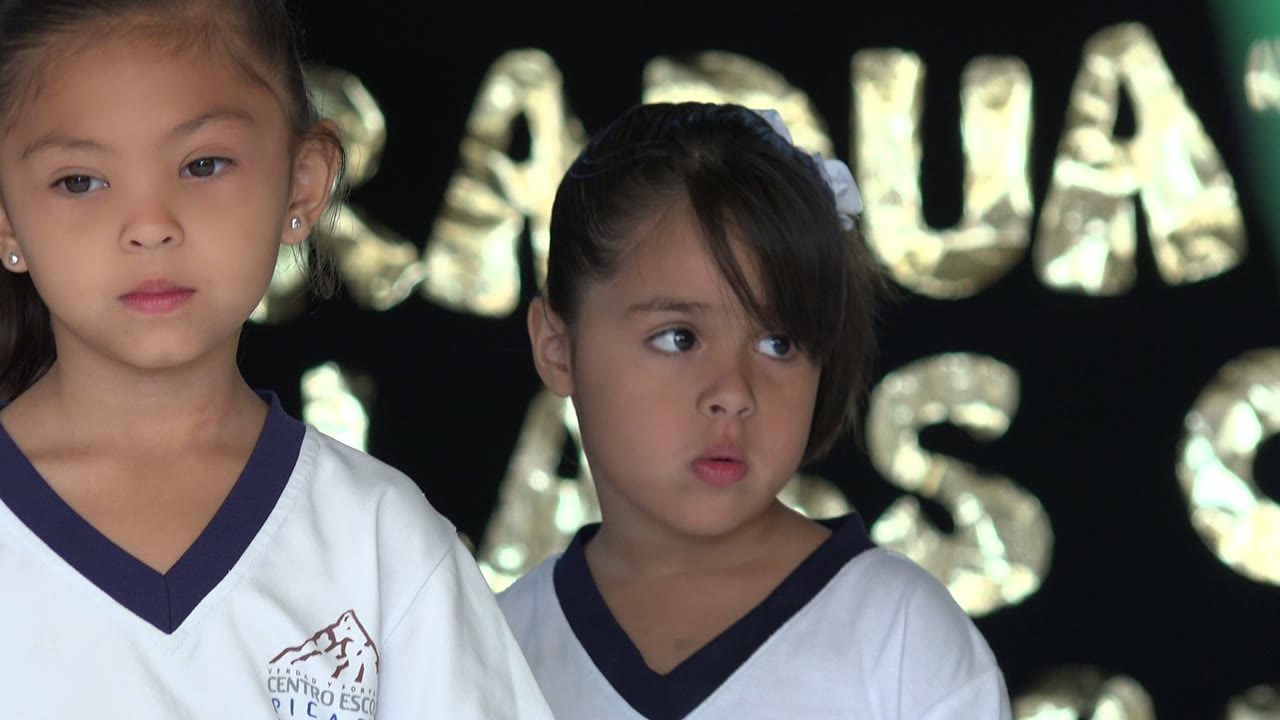 Two girls in white school uniforms in front of gold balloon letters.