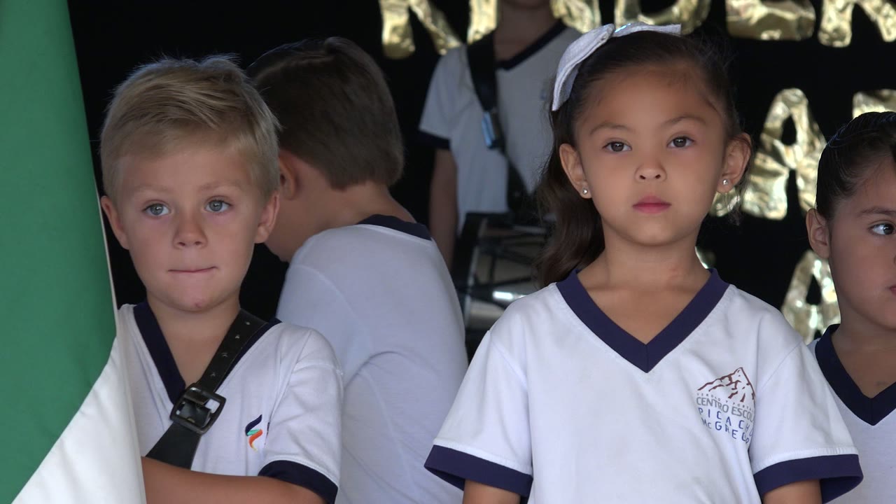 A blond boy and girls in white uniforms stand in San Jose Del Cabo, Mexico, with a green and white flag hanging in the foreground.