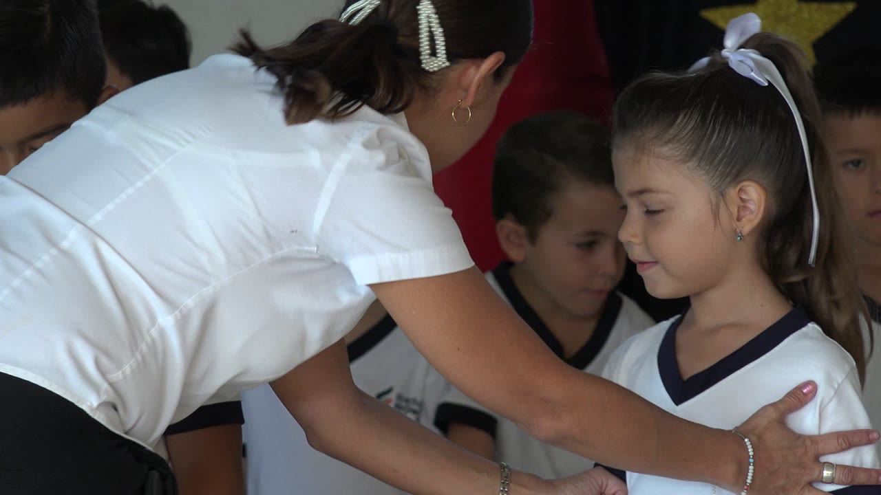 The teacher bends with her back to the camera while several children in white uniforms stand close together beneath gold star decorations in San Jose del Cabo, Mexico.