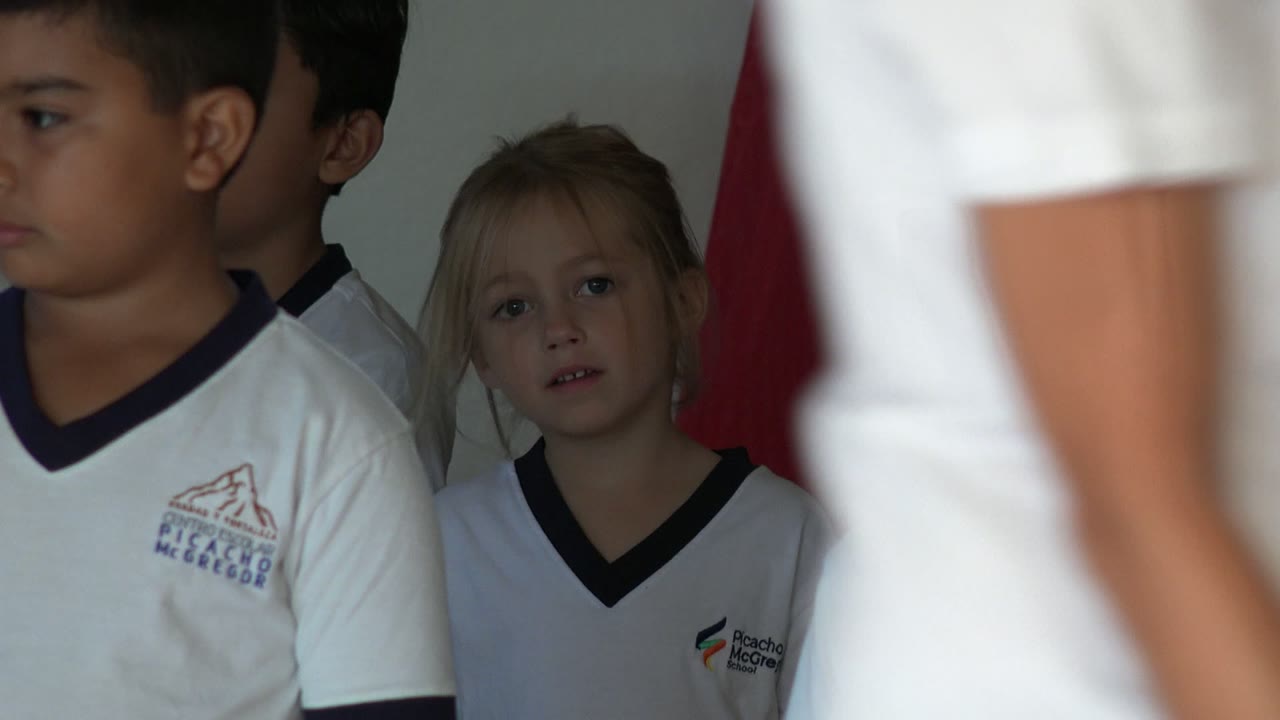 In San Jose del Cabo, Mexico, several children in white shirts stand closely indoors, with a blonde girl near the center and a red vertical object behind them.