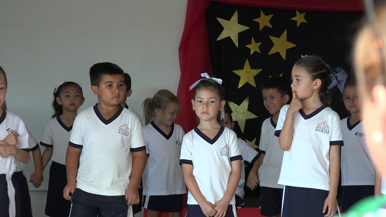 Children in white school uniforms stand before a star backdrop.