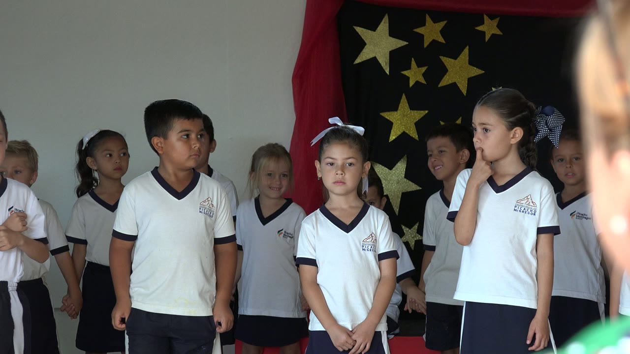 A group of children in matching white uniforms stands in San Jose del Cabo, Mexico, with a central girl facing forward and another girl at right touching her face near the star backdrop.