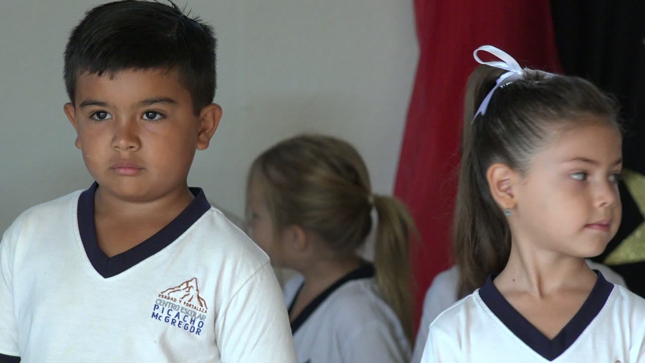 At 0:01 in San Jose Del Cabo, Mexico, a girl with a white bow and a boy in white school shirts stand in the foreground, with a gold star decoration visible between red and black fabric.