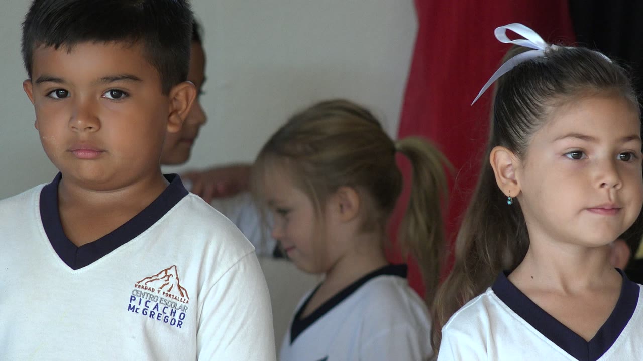 Children in white school uniforms standing indoors near red curtains.