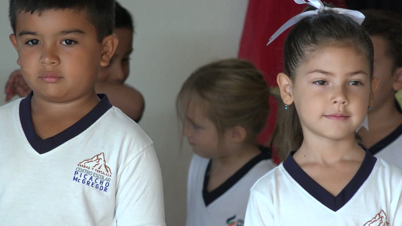 A girl with a white hair bow stands beside a boy in matching white uniforms in San Jose del Cabo, with other children partly visible behind them.