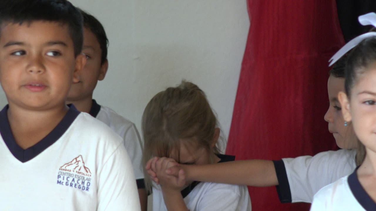 Children in white shirts stand on a small stage near red curtains.