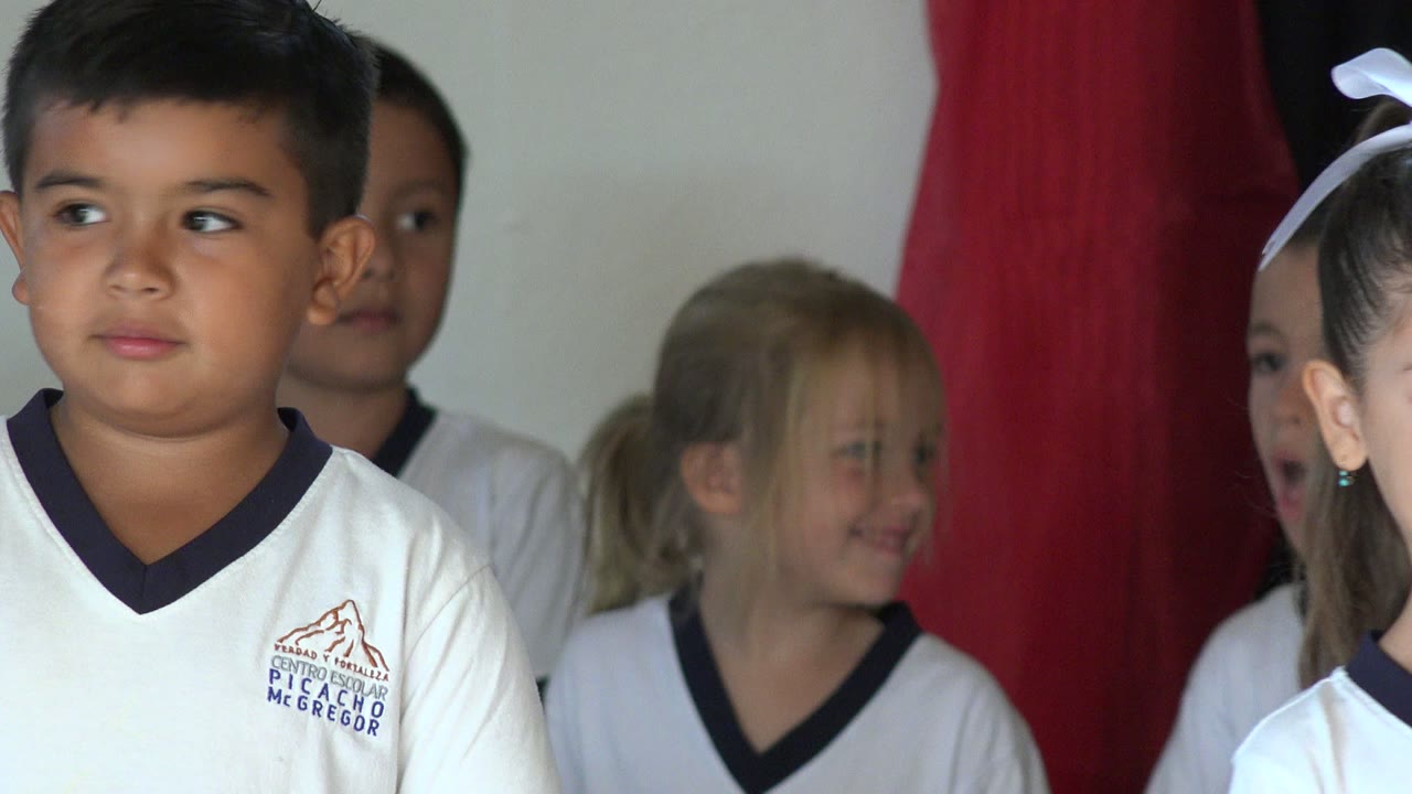 A young boy in a white school shirt faces slightly to the side in San Jose Del Cabo, Mexico, while other children stand behind him near red and dark curtains.