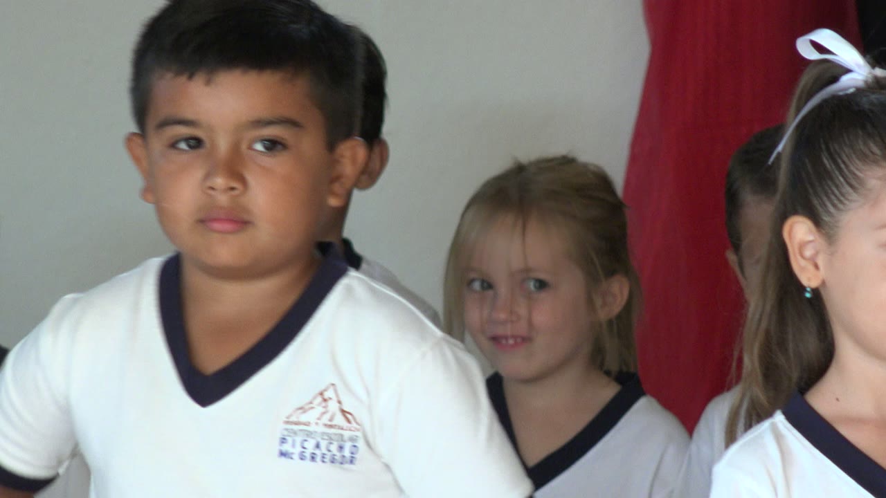 A young boy in a white uniform stands near center in San Jose del Cabo, Mexico, with other children partly visible around him and a red curtain behind the group.