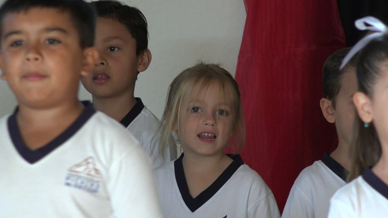 At 0:01, a girl with a white hair bow and a boy in a white shirt stand near the front while other children remain partly visible behind them in San Jose del Cabo, Mexico.