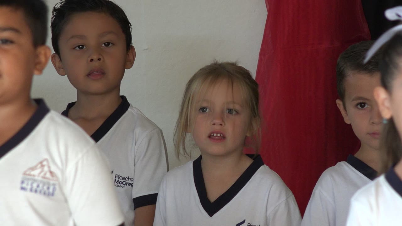 Children in white soccer uniforms stand indoors near a red curtain.