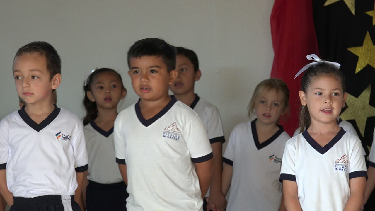 A girl with a white hair bow and boys in white shirts stand among the San Jose del Cabo school choir, with curtain fabric behind them.
