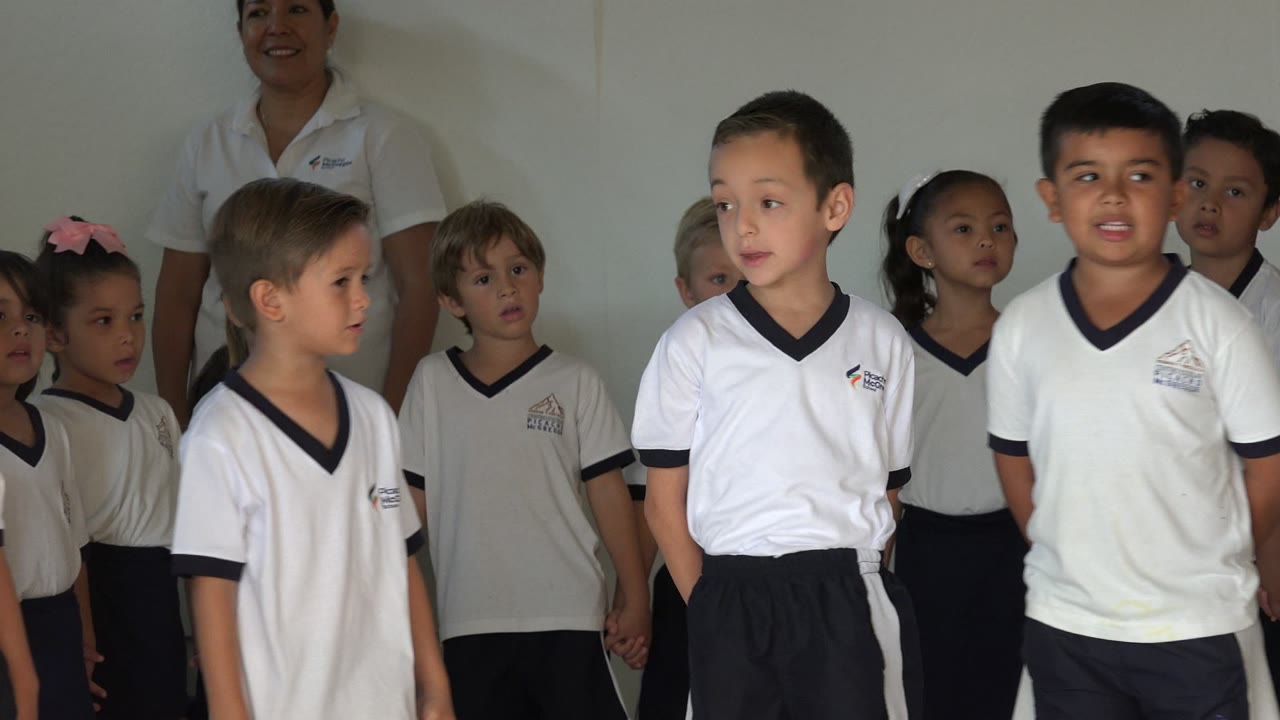 Children in white shirts stand together indoors with an adult behind them.