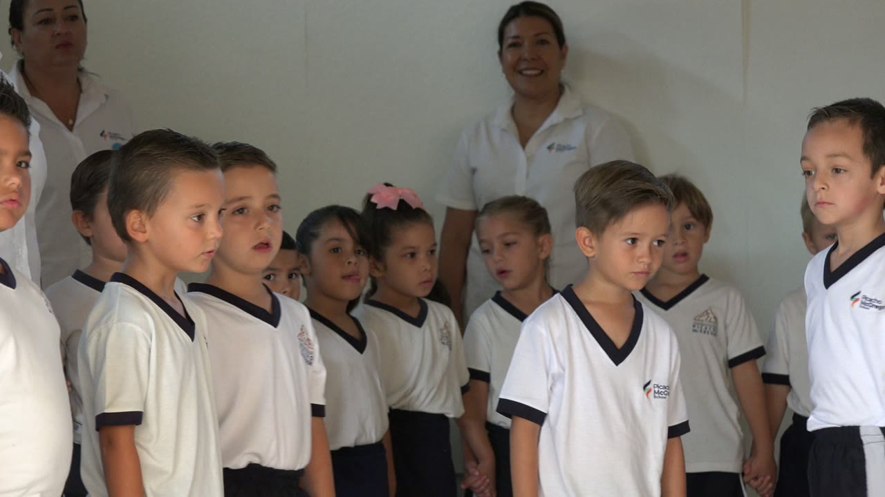 A group of preschool children in San Jose del Cabo faces forward in matching white shirts as an adult supervisor smiles behind the row.
