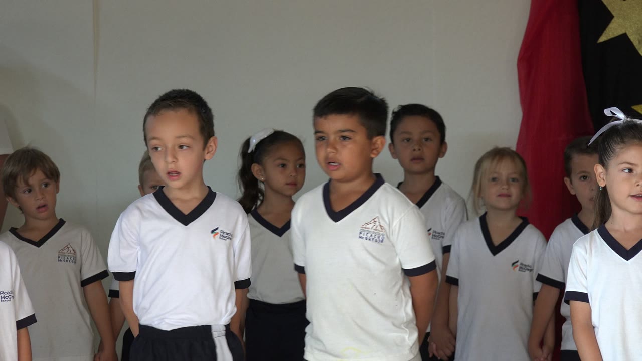 At the start of the San Jose Del Cabo kindergarten performance, several children in white school shirts stand in a row with red fabric and a star backdrop behind them.