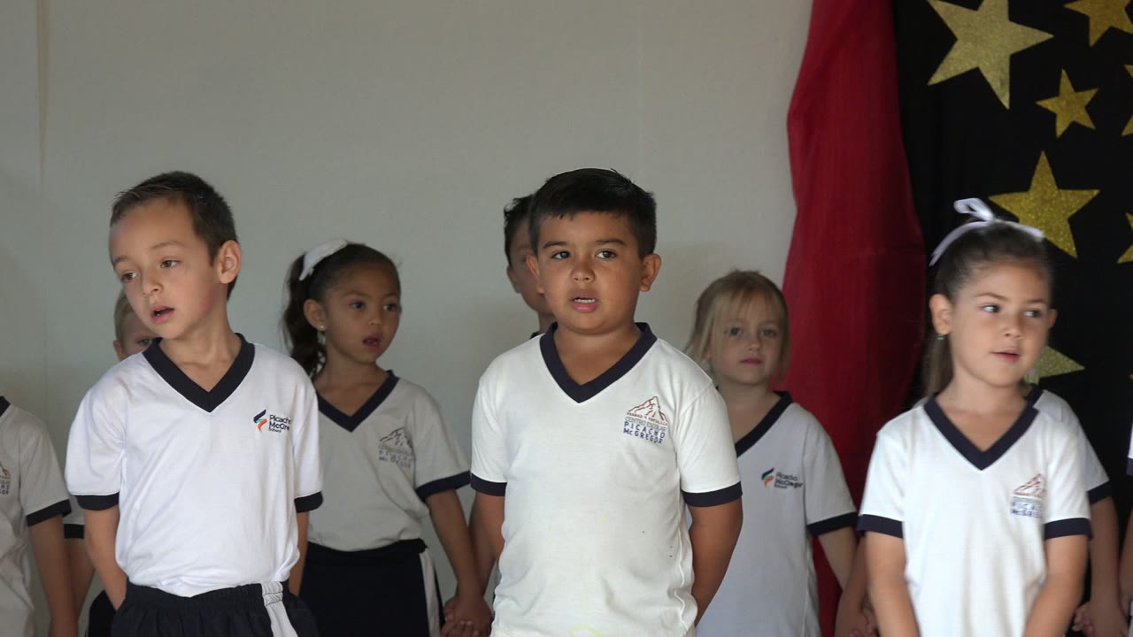 One second in, the children remain grouped on the stage area as a girl with a white hair bow looks sideways and other students face forward or off camera.