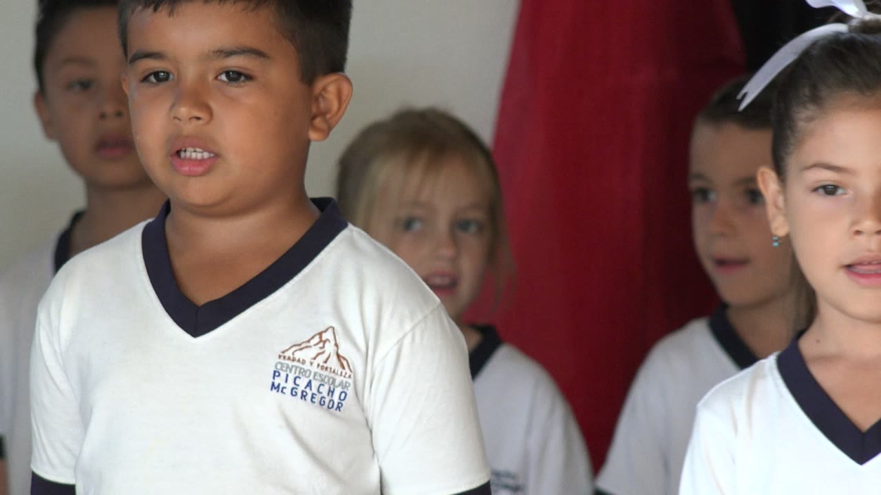 Children in white uniforms singing in front of a red curtain.