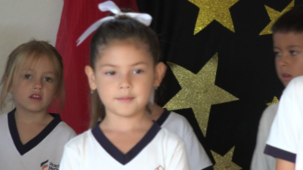 Children in white school shirts stand in front of a star-decorated stage backdrop.