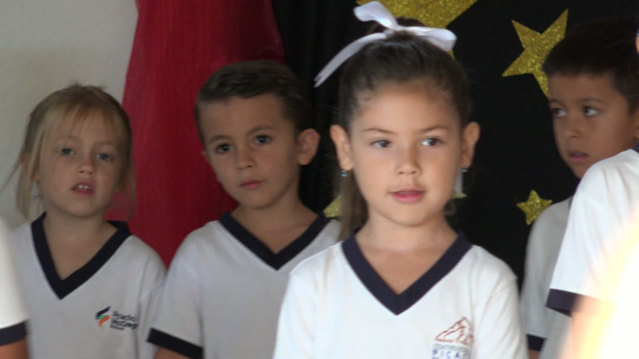 Children in white shirts stand closely together during a San Jose del Cabo school presentation, with red curtains at the sides and gold stars visible behind them.
