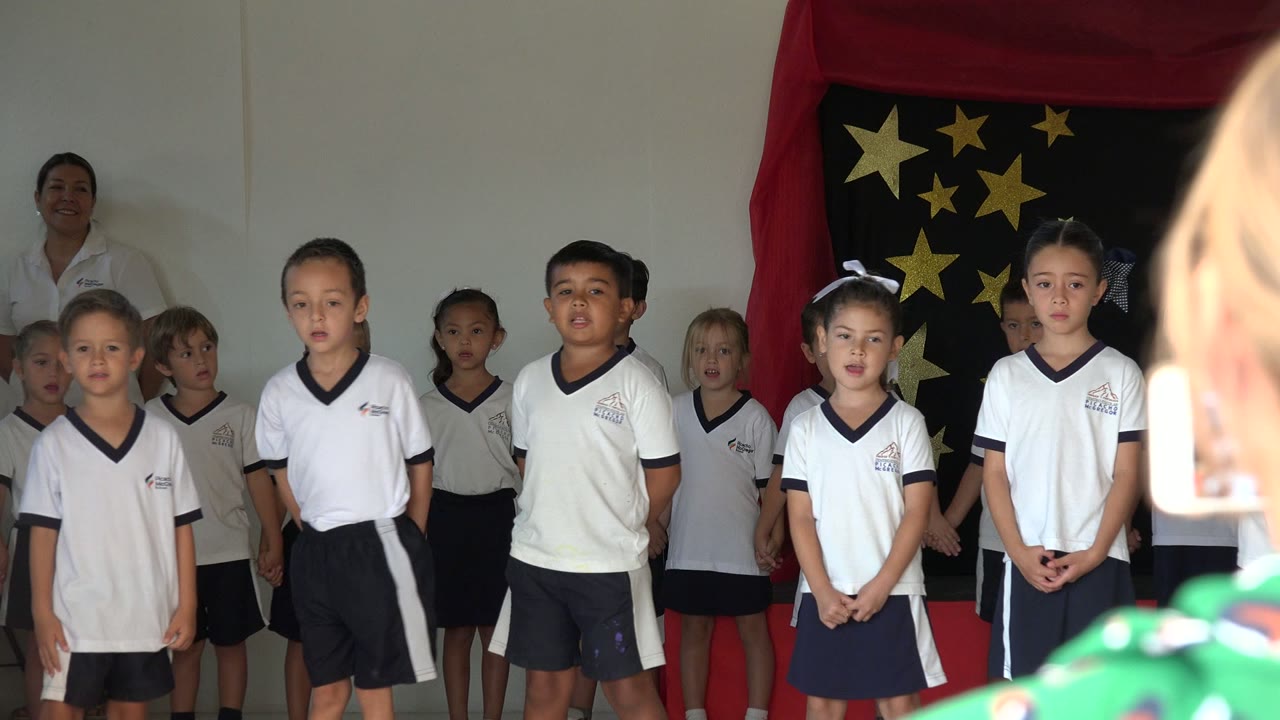 Several children in school uniforms face the camera during the San Jose del Cabo classroom performance, with a red-draped black panel and gold stars behind them.