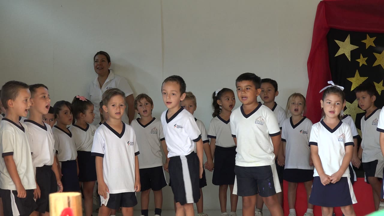 Children in white shirts singing indoors with an adult behind them.