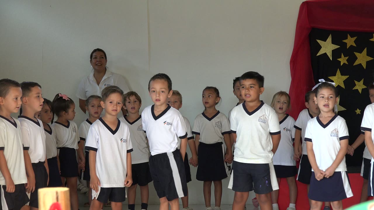 Children in white uniform-like shirts continue singing in San Jose del Cabo, Mexico, with several faces turned forward and the star-decorated backdrop at right.