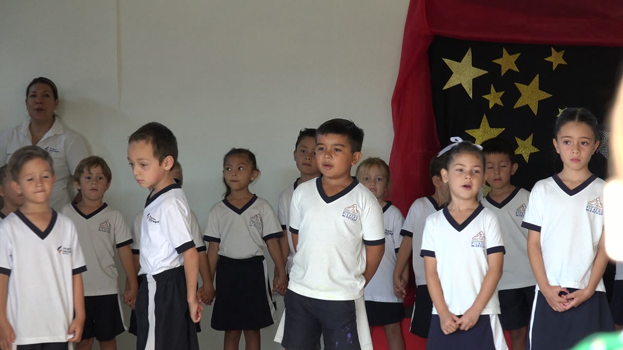 Children in matching white uniforms stand across a small stage in San Jose del Cabo, Mexico, with a teacher at the side and gold stars on a black backdrop.