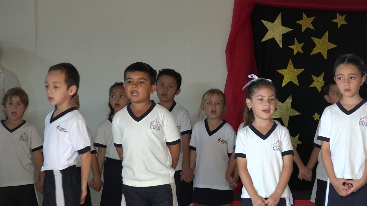 Several children in white shirts and dark bottoms sing on a decorated stage in San Jose del Cabo, Mexico, with red curtains and gold star shapes behind them.
