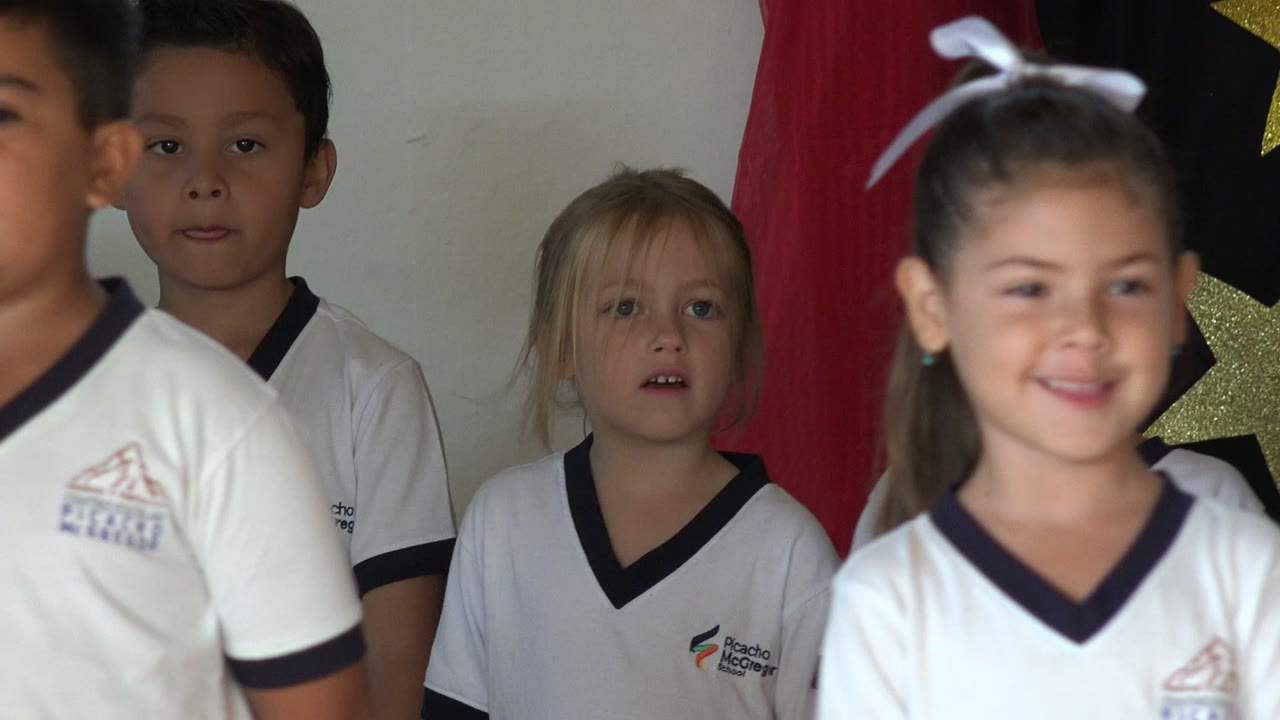 Young children in white school uniforms sing during a graduation ceremony in San Jose del Cabo, Mexico, with red drapes and star decorations behind them.