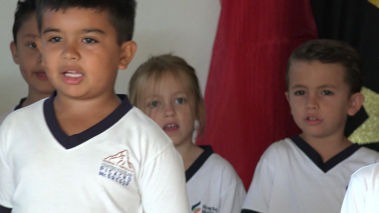 A girl with a white hair bow and a boy in a white uniform shirt stand near the camera while classmates sing behind them in San Jose del Cabo, Mexico.
