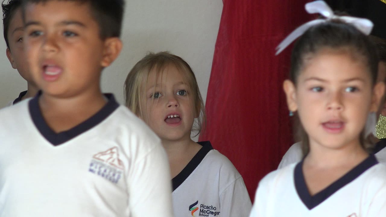 Children in white school shirts singing indoors near a red backdrop.
