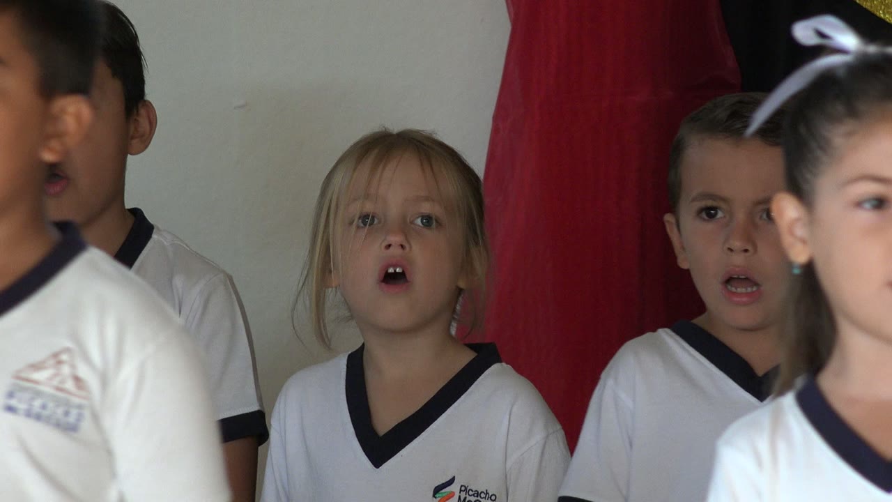 A girl with a white hair bow moves through the foreground while other children in white school shirts sing behind her at the San Jose del Cabo concert.