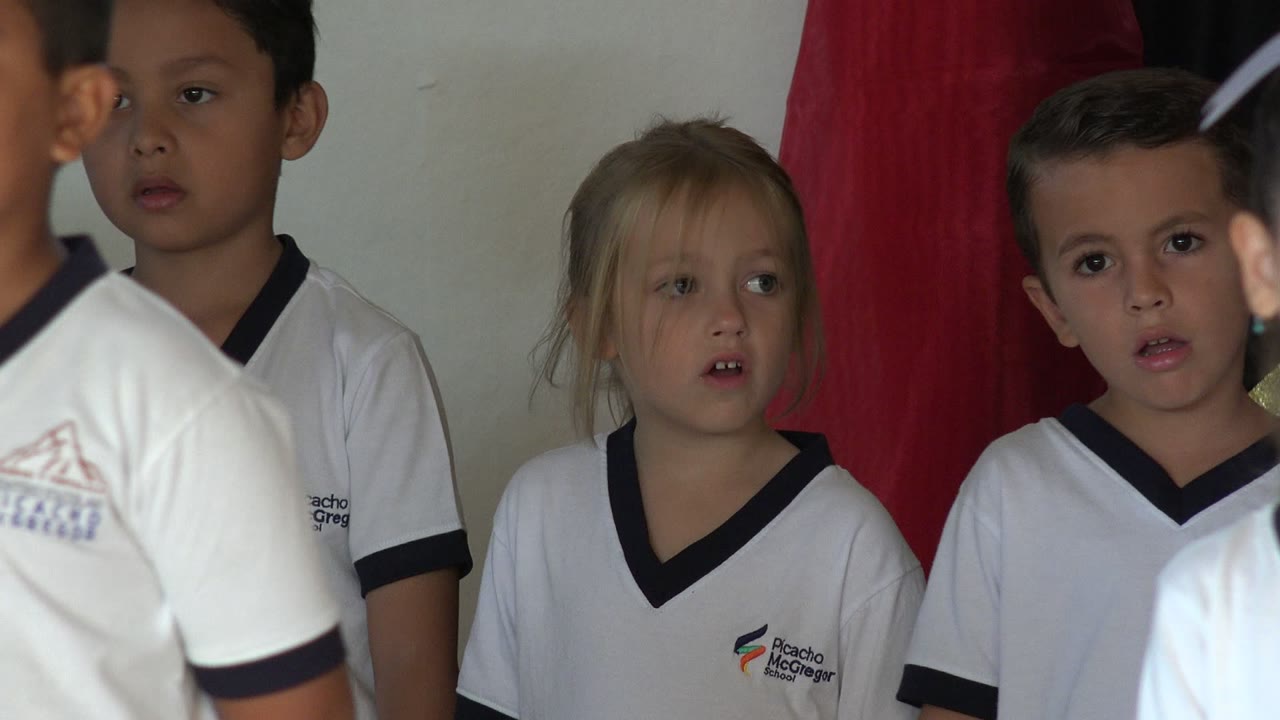 Young children in white school uniforms stand close together in San Jose del Cabo, Mexico, with a girl and boy visible near a red fabric backdrop.