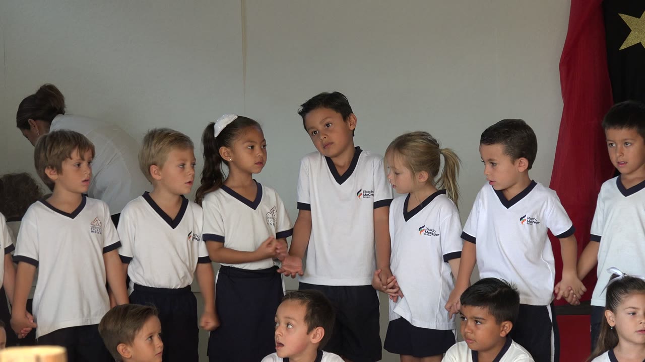 Children in white uniforms stand and sit in a classroom beside a red and black flag.