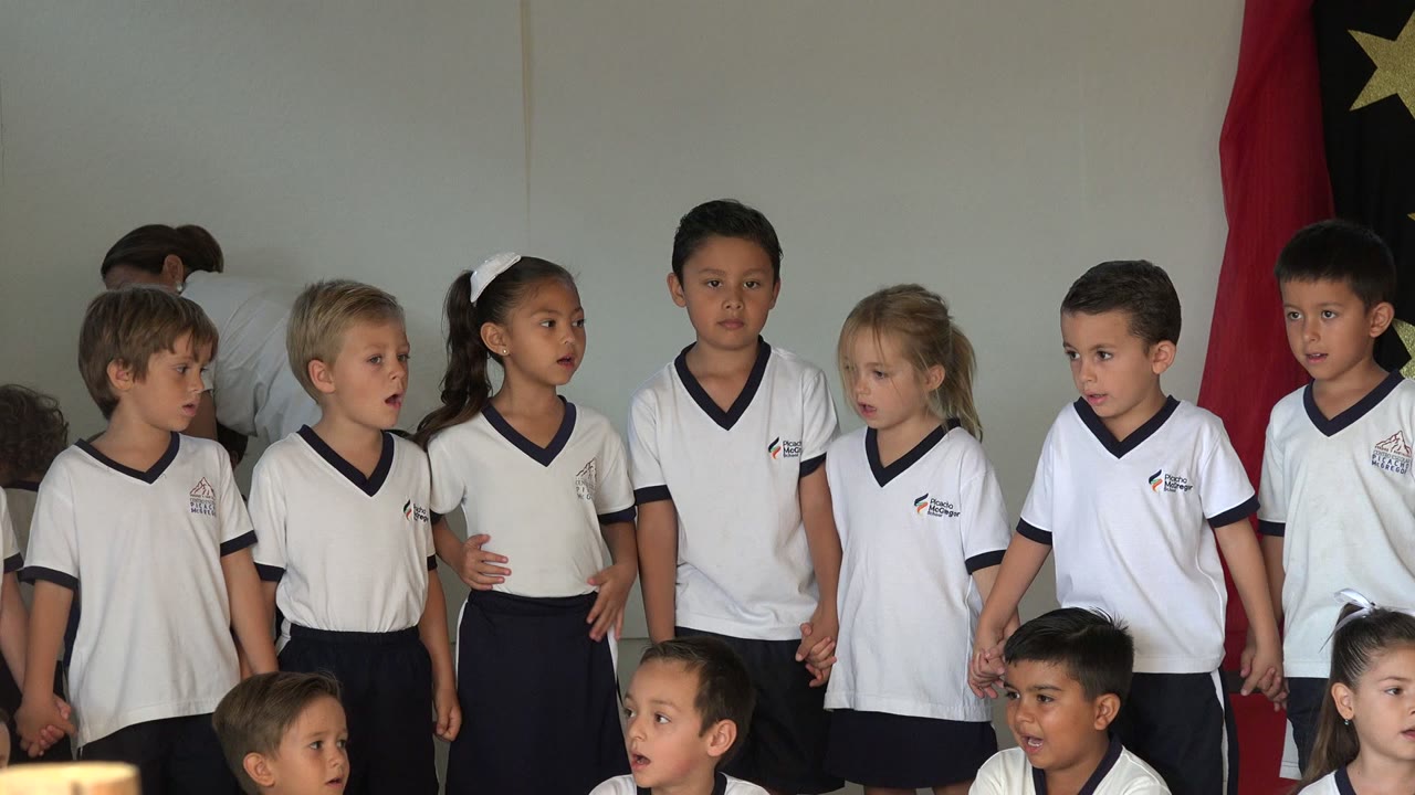 At 0:01, a row of young students in white shirts and dark shorts or skirts stands in the classroom in San Jose del Cabo, Mexico, while seated classmates are partly visible below.