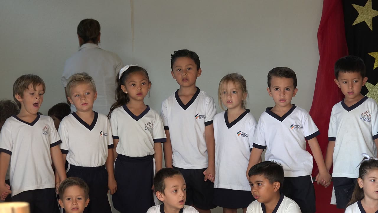 Primary school children in San Jose Del Cabo stand in white uniforms inside a classroom while an adult teacher faces away near the back wall.