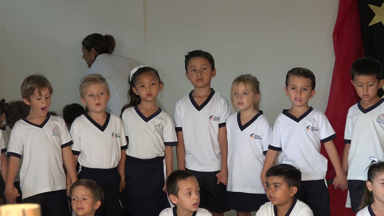 A row of primary school children in San Jose Del Cabo faces forward in white shirts as a red flag with yellow stars hangs behind them.