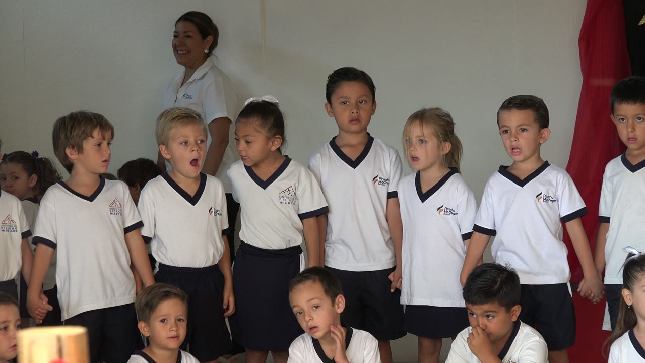 Children in white uniforms stand in a row with a smiling adult behind them.