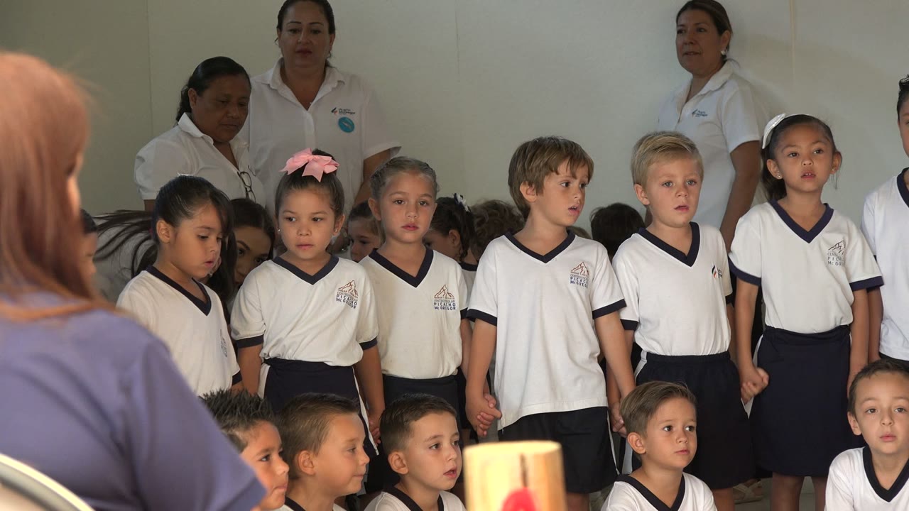 At 0:00, children in white shirts with navy trim stand in rows in San Jose del Cabo, Mexico, with adult staff behind them and audience figures at the foreground edge.