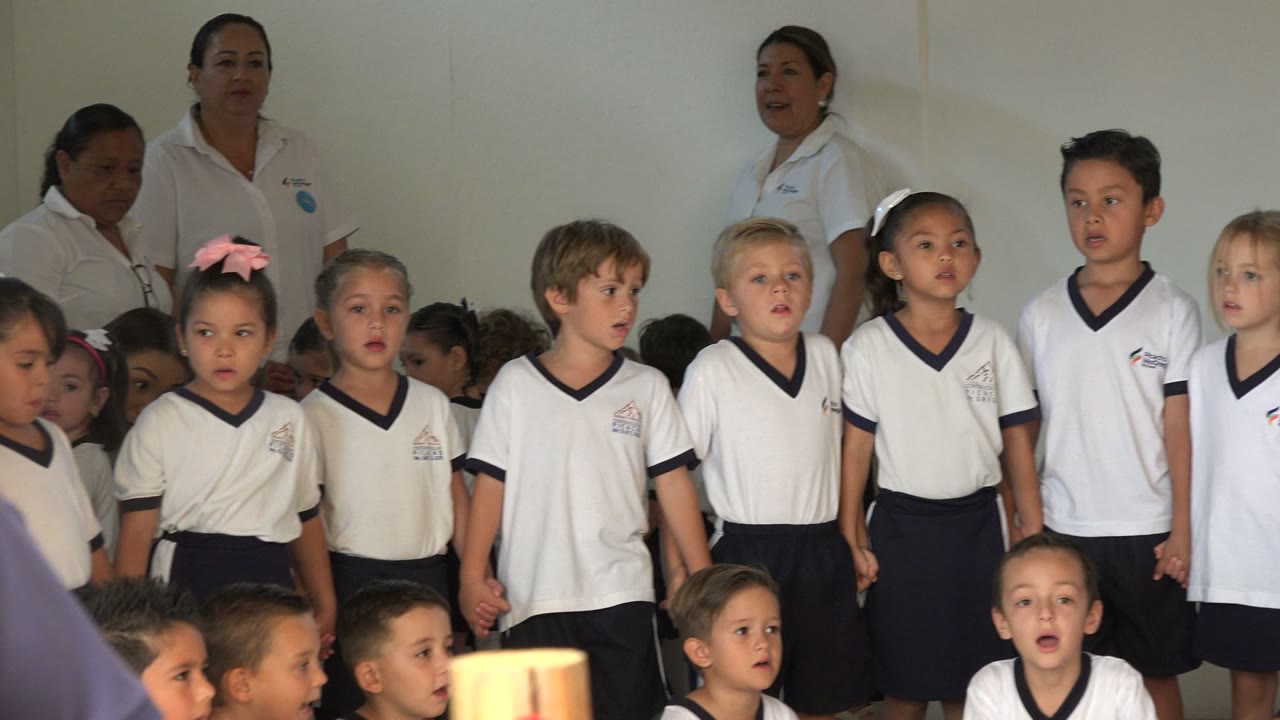 At 0:01, the line of children continues across the frame in San Jose del Cabo, Mexico, with several faces turned toward the front and adults standing against the pale wall.