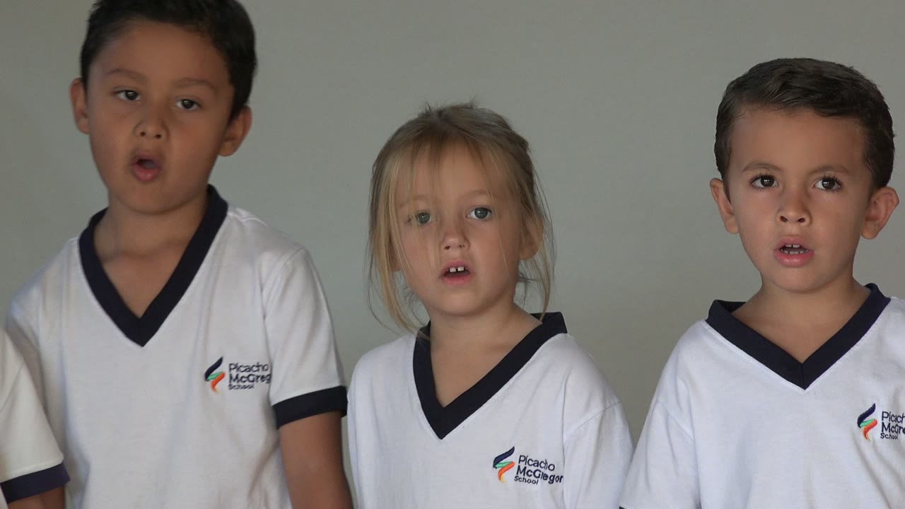 Three children in matching white uniforms are framed closely in San Jose del Cabo, Mexico, with two open mouths suggesting the group singing continues.