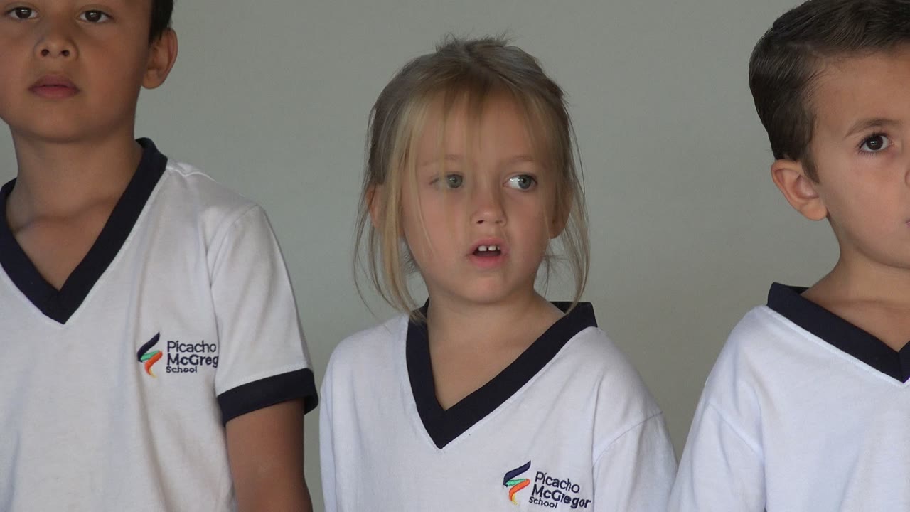 Young children in white school uniforms standing against a plain wall.