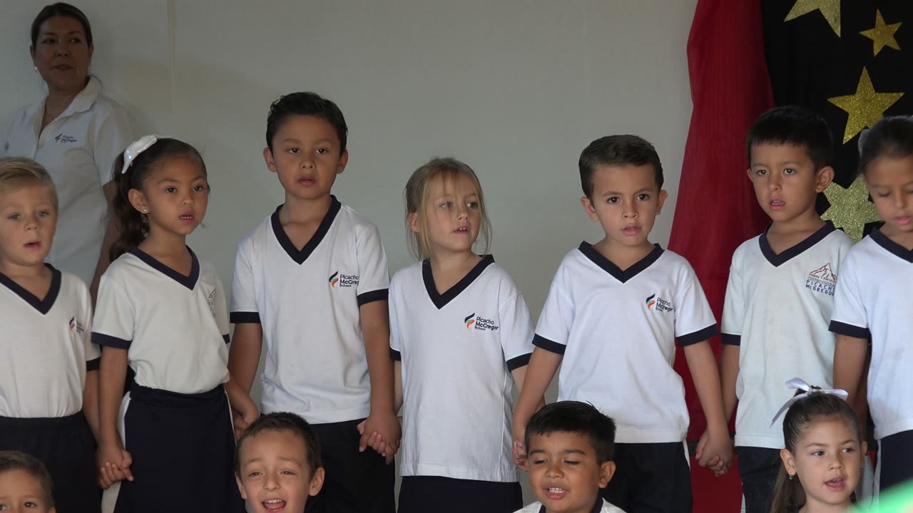 Children in white shirts and dark bottoms stand together in San Jose del Cabo, Mexico, with adult staff partly visible behind them and red and black star fabric in the background.