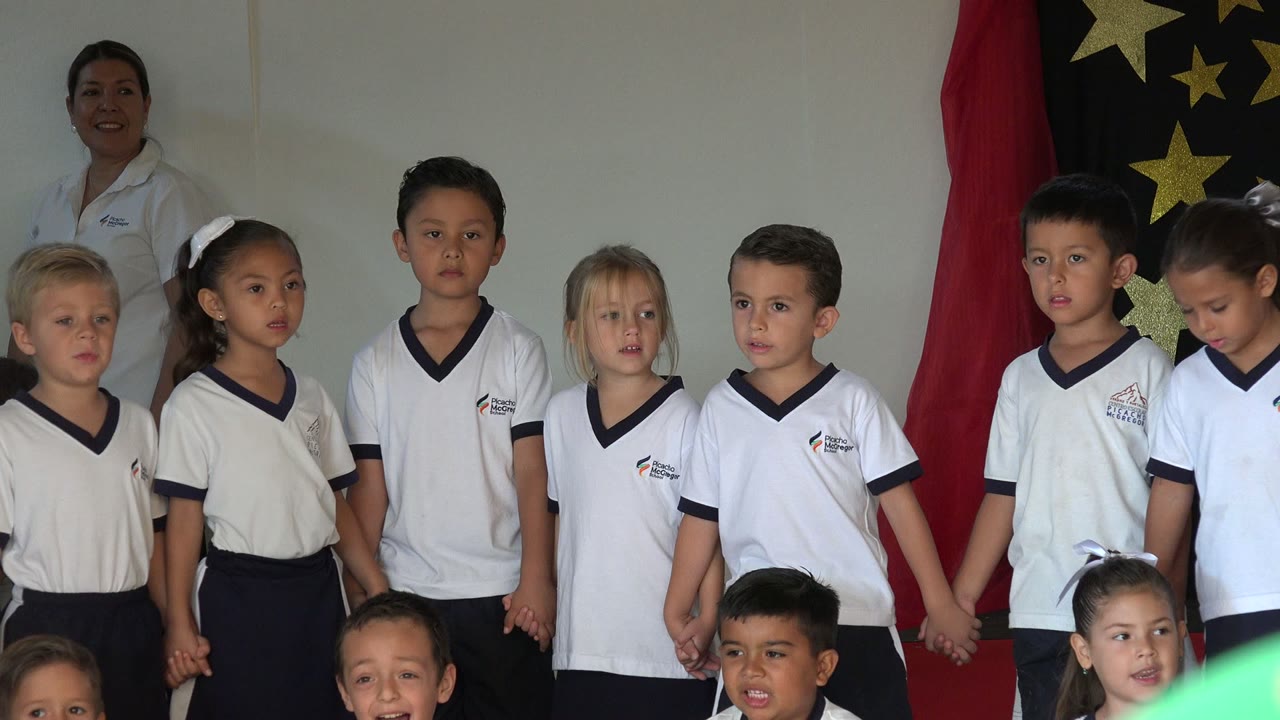 A row of children in white uniform-style shirts stands hand in hand in San Jose del Cabo, Mexico, while a black fabric panel with gold stars hangs behind them.