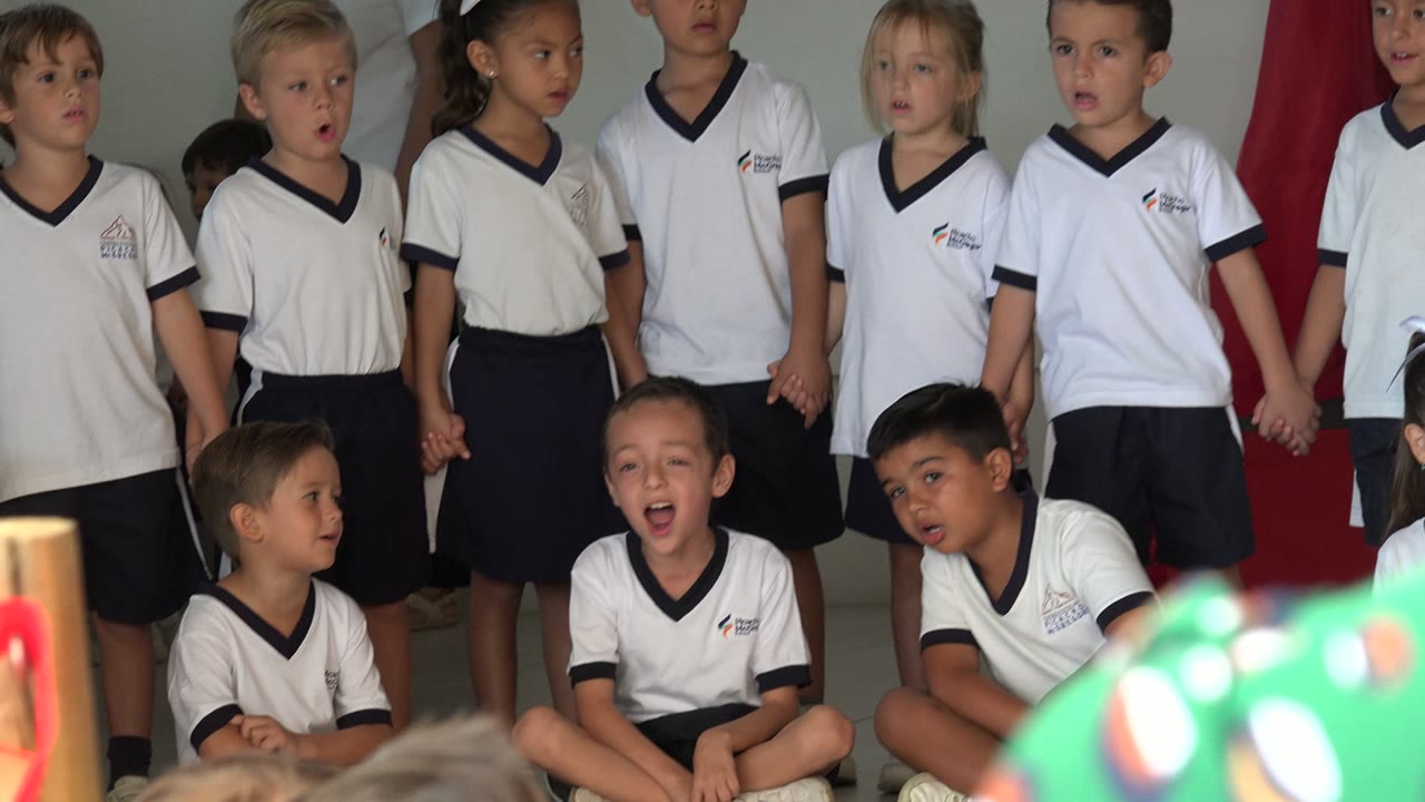 Children in matching white shirts and dark shorts sing in a group indoors in San Jose del Cabo, Mexico, with audience heads partly visible at the bottom of the frame.