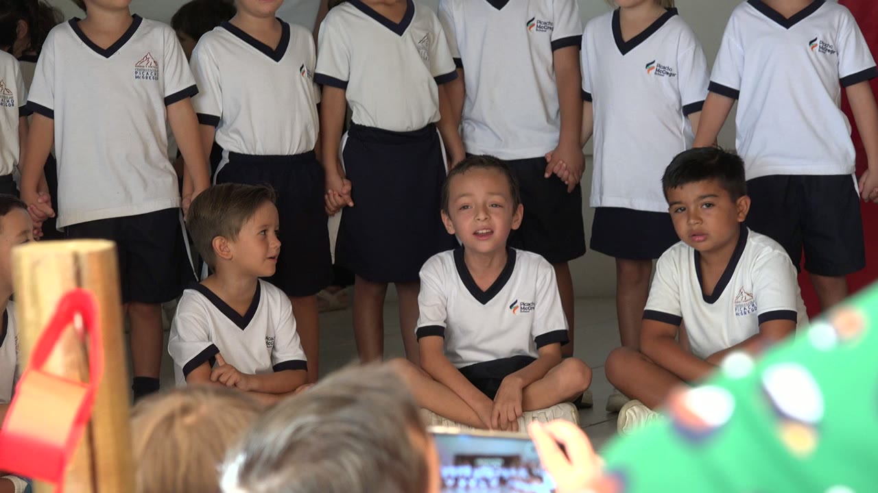 A row of children in white uniform shirts stands behind seated classmates during the choir performance in San Jose del Cabo, Mexico, with colorful foreground objects partly blocking the lower frame.