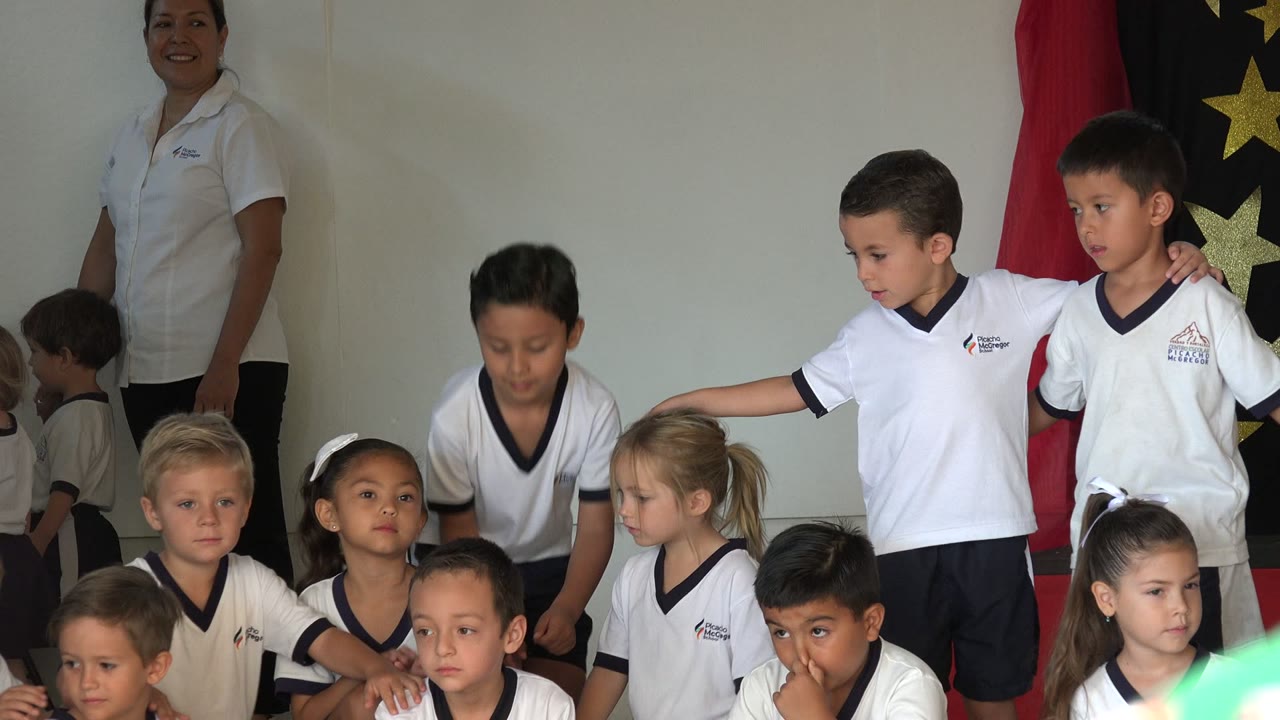 Children in white school shirts gather with a teacher standing behind them.
